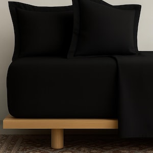May include: A black bed set, comprising a fitted sheet and two pillowcases, is presented on a light wood bed frame. The pillowcases feature a contrasting border. The bed is against a neutral wall, with a patterned rug visible.