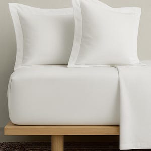 May include: A crisp, white bedding set featuring two pillowcases and a fitted sheet. The pillowcases have a tailored edge, and the sheets are neatly arranged on a wooden bed frame. The overall aesthetic is clean and minimalist.