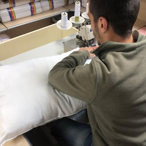 May include: A person sewing a white pillow on a sewing machine. The person is wearing a green sweater and blue jeans. There are spools of thread on the machine.