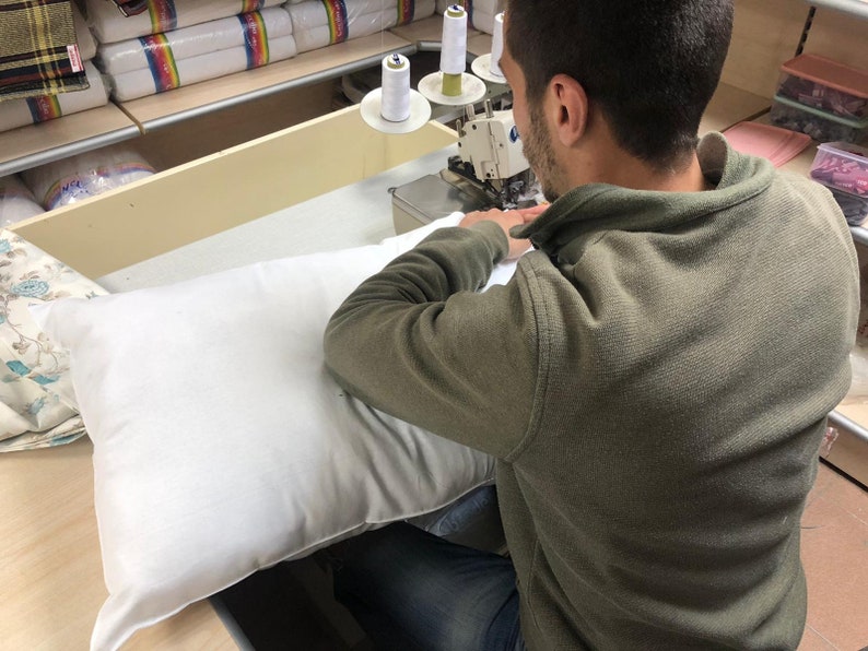 May include: A person sewing a white pillow on a sewing machine. The person is wearing a green sweater and blue jeans. There are spools of thread on the machine.