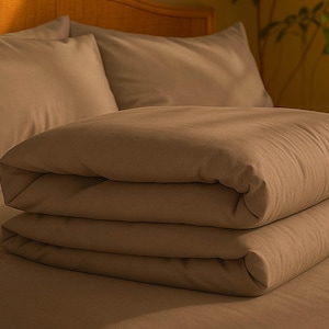 May include: A bed with a folded, stacked comforter and two pillows. The bedding is a solid, warm brown color. The pillows are behind the comforter, and the bed frame is visible in the background.