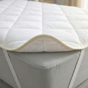 May include: A white quilted mattress topper with a cream-colored border is shown on a bed. The topper is secured with elastic straps. The bed's gray fitted sheet is visible beneath the topper. The topper has a diamond pattern.