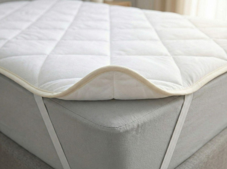 Puede incluir: Un protector de colch&oacute;n acolchado blanco con una textura suave y un borde ribeteado en color beige claro. El protector presenta un patr&oacute;n de costuras en cuadr&iacute;cula y se sujeta a un colch&oacute;n gris con correas el&aacute;sticas blancas en las esquinas.