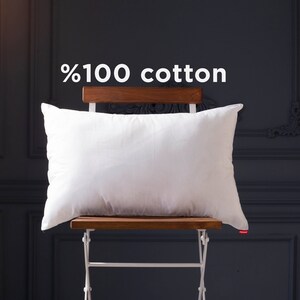 May include: A white pillow made of 100% cotton, sitting on a wooden chair with a white metal frame against a dark gray wall.