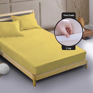 Può includere: Un lenzuolo con angoli elasticizzato giallo con un elastico spesso intorno al bordo. L'elastico è spesso 4 cm. Il lenzuolo è su un letto con un cuscino giallo e bianco.