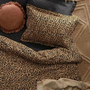May include: A bed with black sheets, a leopard print duvet cover and pillowcases. A round, fringed, peach-colored pillow is on the bed. Dried ornamental grass and a beige rug are in the background.