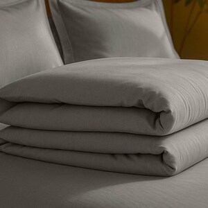 May include: A folded, light gray duvet cover and pillowcases on a bed. The bedding is neatly stacked, showcasing a smooth, soft texture. The neutral color palette creates a calming and minimalist aesthetic, suitable for bedroom decor.