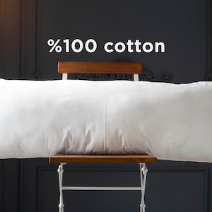 May include: A white, rectangular pillow made of 100% cotton, resting on a wooden chair with white legs. The pillow is sitting on a dark gray background.