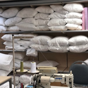 May include: A white industrial sewing machine with two spools of thread, one green and one white, on a table with a stack of white pillows in the background. The machine has a JUKI label.