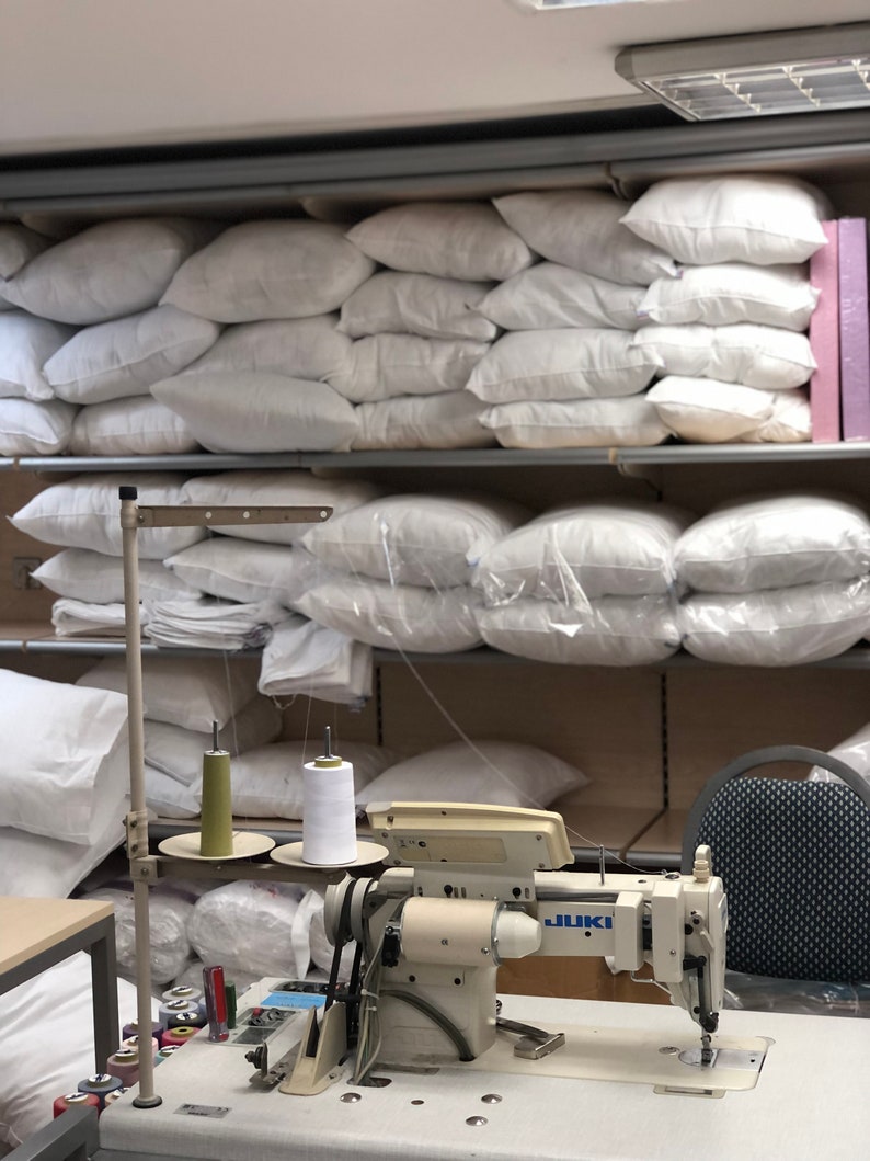 May include: A white industrial sewing machine with two spools of thread, one green and one white, on a table with a stack of white pillows in the background. The machine has a JUKI label.