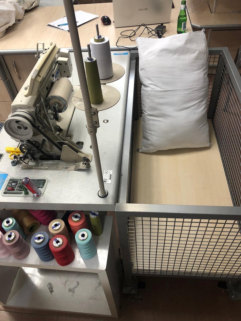May include: A white sewing machine with a metal frame and a white pillow on a wooden table. The machine has a spool of thread and a needle. There are also spools of thread in different colors on a shelf below the machine.