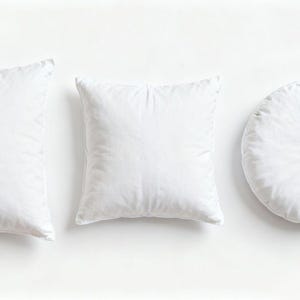 Puede incluir: Tres almohadas blancas de diferentes formas: rectangular, cuadrada y redonda. Las almohadas son lisas y parecen estar rellenas de un material suave. Están dispuestas sobre una superficie blanca.
