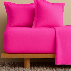 Op de afbeelding: Een felroze beddengoedset met twee kussenslopen en een hoeslaken. Het beddengoed wordt gepresenteerd op een houten platform tegen een neutrale achtergrond. De set is effen van kleur en biedt een moderne en minimalistische uitstraling.