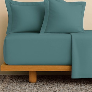 Pode incluir: Conjunto de lençóis e fronhas de cor azul-petróleo, exibidos numa estrutura de cama de madeira. O conjunto de roupa de cama inclui um lençol com elástico e duas fronhas com um design simples. Os lençóis estão bem arrumados, mostrando a textura suave.