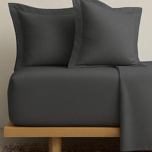Peut inclure: Ensemble de linge de lit gris foncé, comprenant deux taies d'oreiller et un drap-housse, présenté sur un cadre de lit en bois. Les taies d'oreiller ont un bord subtil. L'esthétique générale est minimaliste et moderne, adaptée à la décoration de chambre.