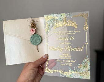 以下が含まれることがあります： 金色の文字と花のアクセントが施されたSweet 16の招待状。招待状は開かれており、ワックスシールと装飾が施された封筒が現れます。招待状には、両親の名前、娘の名前、イベントの詳細が含まれています。