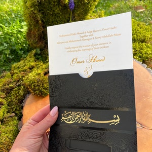 May include: A white wedding invitation card with gold lettering announcing the marriage of Omar Ahmed and Asiya Hussein Omar-Hashi. The card is decorated with black and gold floral patterns.