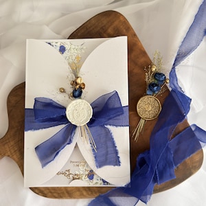 Puede incluir: Una invitación blanca con una cinta azul y un sello de cera. La invitación tiene un diseño floral y el texto "Presentation 7:30 PM". También hay un pequeño arreglo floral y una moneda de oro.