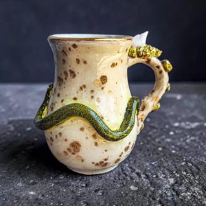 Könnte beinhalten: Ein Keramikbecher mit cremefarbener Basis und braunen Sprenkeln. Eine texturierte, grün-gelbe schlangenartige Form windet sich um den Körper. Der Henkel ist wie ein Ast geformt, mit kleinen gelben Auswüchsen und einem weißen kristallartigen Akzent.