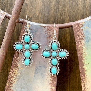 May include: A pair of silver-toned cross earrings. Each cross features oval turquoise-colored stones set within a detailed silver frame. The earrings hang from simple silver hooks, showcasing a Southwestern or Western style.