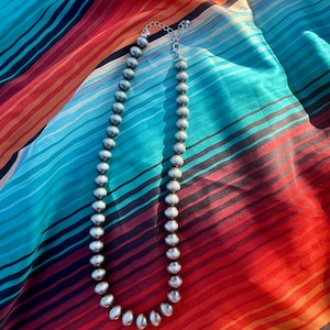Pu&ograve; includere: Una collana di perline d'argento con una delicata chiusura a catena poggia su un tessuto a righe colorato. La collana presenta perline rotonde e uniformemente distanziate. Il tessuto ha un vivace motivo a righe rosse, arancioni, blu e nere.