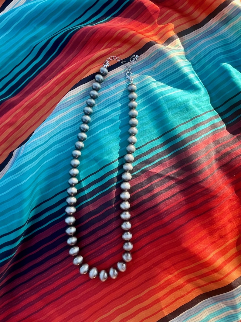 Pu&ograve; includere: Una collana di perline d'argento con una delicata chiusura a catena poggia su un tessuto a righe colorato. La collana presenta perline rotonde e uniformemente distanziate. Il tessuto ha un vivace motivo a righe rosse, arancioni, blu e nere.