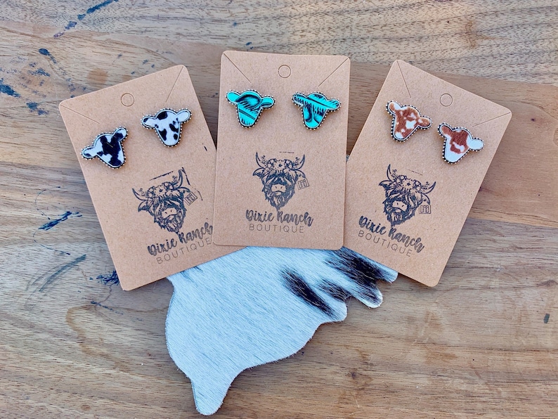 May include: Three pairs of cowhide earrings with silver posts. The earrings are in the shapes of cow heads with different color patterns. The earrings are displayed on brown cards with the text "Dixie Ranch Boutique".