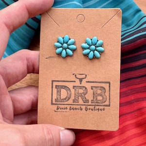 May include: Turquoise flower stud earrings displayed on a brown card with the Dixie Ranch Boutique logo. The earrings are held by a hand, with a colourful striped fabric in the background.