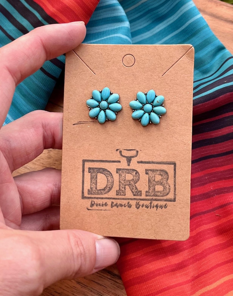 May include: Turquoise flower stud earrings displayed on a brown card with the Dixie Ranch Boutique logo. The earrings are held by a hand, with a colourful striped fabric in the background.