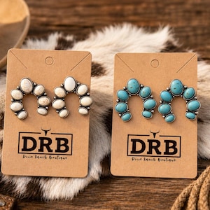 May include: Two pairs of horseshoe-shaped earrings on display. One pair features white stones, the other turquoise. Each earring set is mounted on a brown card with the "DRB Dixie Ranch Boutique" logo. The background includes a cowboy hat and a cactus.