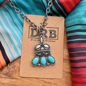Puede incluir: Collar plateado con un colgante con tres piedras de color turquesa. El colgante tiene un dise&ntilde;o ornamentado con forma rectangular y una parte superior decorativa. El collar se muestra en una tarjeta marr&oacute;n con el texto "DRB Dixie Rose Boutique".