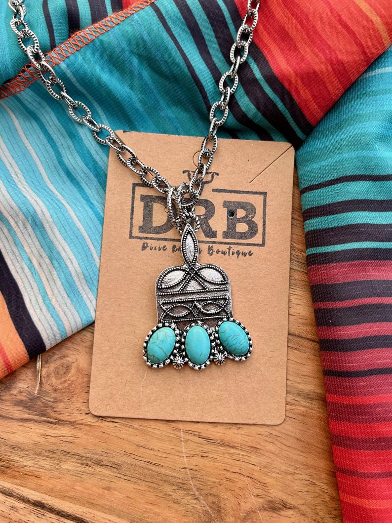 Puede incluir: Collar plateado con un colgante con tres piedras de color turquesa. El colgante tiene un dise&ntilde;o ornamentado con forma rectangular y una parte superior decorativa. El collar se muestra en una tarjeta marr&oacute;n con el texto "DRB Dixie Rose Boutique".