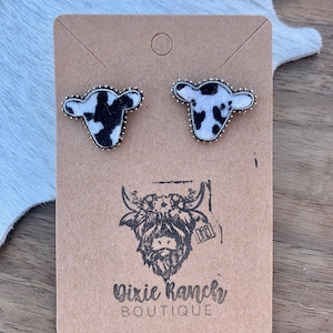 May include: A pair of cowhide earrings with silver accents. The earrings are shaped like a cow's head and have a black and white cowhide pattern. The earrings are on a brown card with the text "Dixie Ranch Boutique" and a logo of a cow's head.