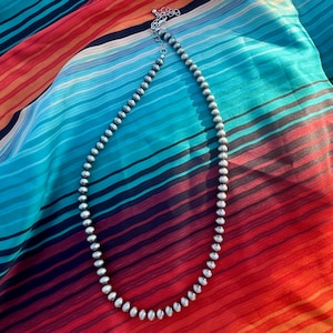 Pu&ograve; includere: Una collana di perline d'argento con chiusura a moschettone &egrave; appoggiata su un tessuto a righe colorato. La collana presenta piccole perline rotonde. Il tessuto ha un vivace motivo a righe rosse, arancioni, blu e nere.