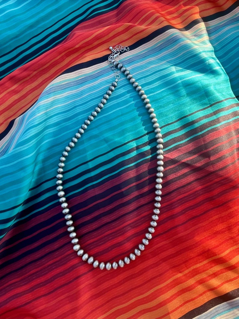 Pu&ograve; includere: Una collana di perline d'argento con chiusura a moschettone &egrave; appoggiata su un tessuto a righe colorato. La collana presenta piccole perline rotonde. Il tessuto ha un vivace motivo a righe rosse, arancioni, blu e nere.