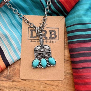 Puede incluir: Collar plateado con un colgante con tres piedras turquesas ovaladas. El colgante tiene un dise&ntilde;o ornamentado y est&aacute; unido a una cadena. El collar descansa sobre una etiqueta marr&oacute;n con el texto "DRB Dusee Rd Boutique". El fondo incluye tela a rayas coloridas.