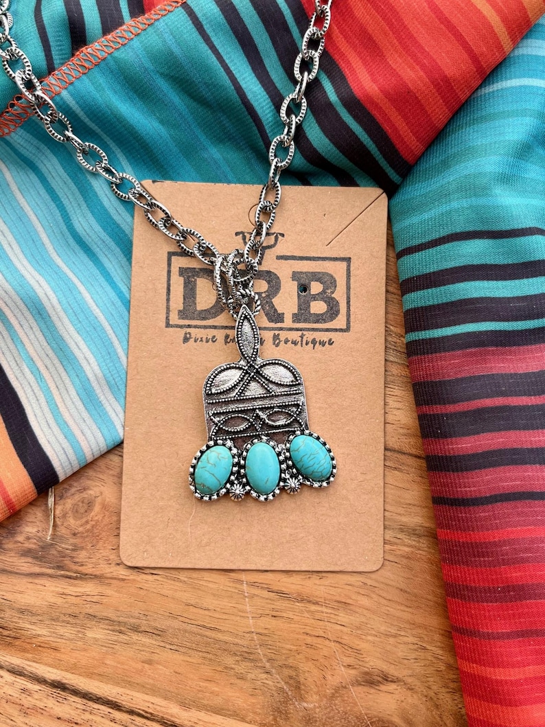 Puede incluir: Collar plateado con un colgante con tres piedras turquesas ovaladas. El colgante tiene un dise&ntilde;o ornamentado y est&aacute; unido a una cadena. El collar descansa sobre una etiqueta marr&oacute;n con el texto "DRB Dusee Rd Boutique". El fondo incluye tela a rayas coloridas.