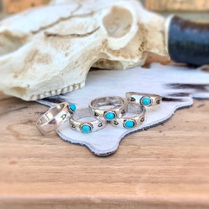 May include: A collection of silver-toned rings with turquoise-colored stones. Each ring features a band with a decorative pattern. The rings are displayed on a white leather surface, with a cow skull in the background.