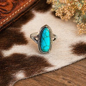May include: Silver ring with an oval turquoise stone. The ring has a detailed silver setting and a split band design. The turquoise stone is a vibrant blue-green color, set against a neutral background.