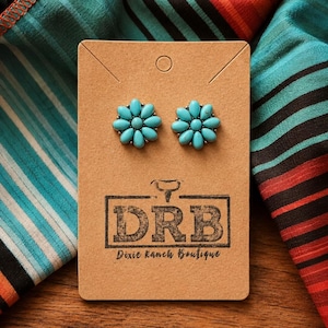 May include: Turquoise flower stud earrings presented on a brown card, with the text "DRB Dixie Ranch Boutique". The earrings are set against a backdrop of patterned fabric in various colours.