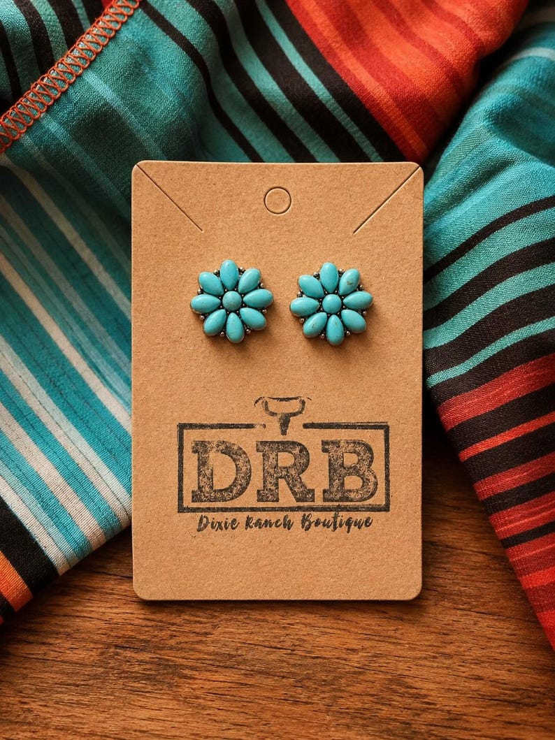 May include: Turquoise flower stud earrings presented on a brown card, with the text "DRB Dixie Ranch Boutique". The earrings are set against a backdrop of patterned fabric in various colours.