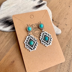 May include: A pair of silver earrings with turquoise stones and a geometric design. The earrings are on a brown card with a white circle in the top right corner.