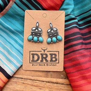 May include: Silver-toned earrings with turquoise-colored stones. The earrings are displayed on a brown card with the text "DRB Dixie Ranch Boutique" and are set against a colorful striped fabric.