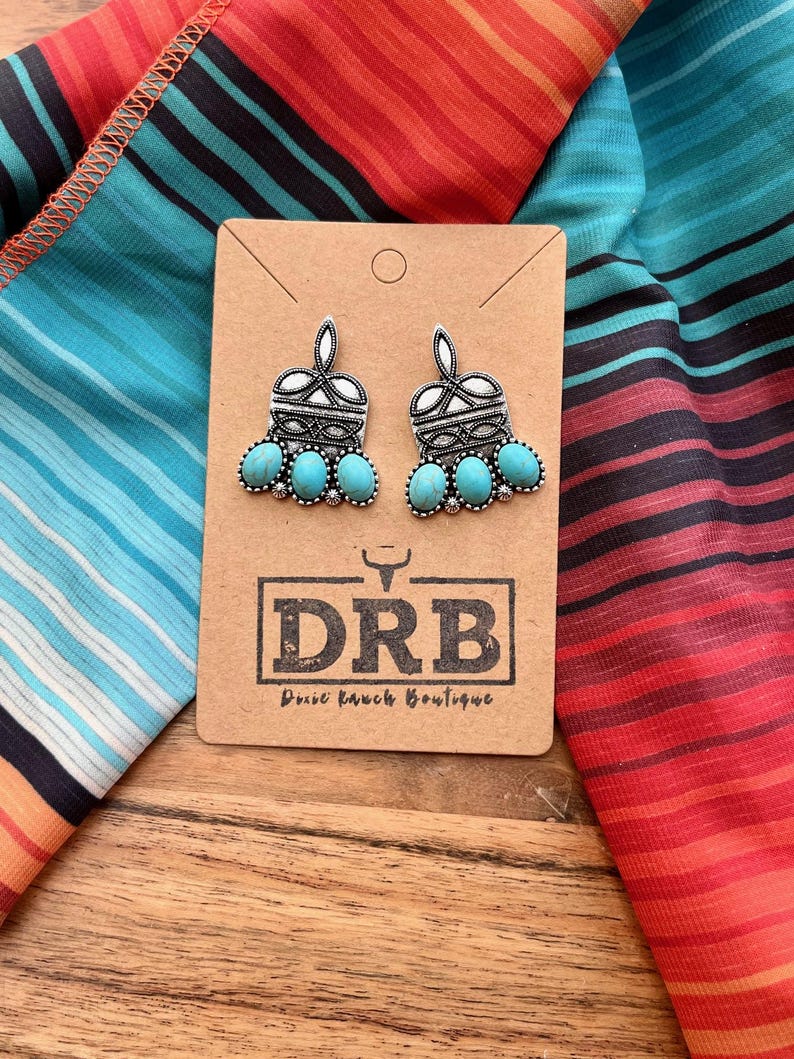 May include: Silver-toned earrings with turquoise-colored stones. The earrings are displayed on a brown card with the text "DRB Dixie Ranch Boutique" and are set against a colorful striped fabric.
