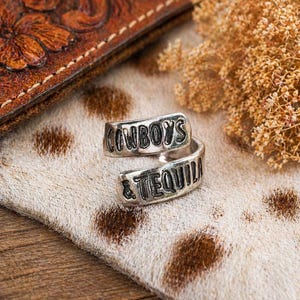 May include: A silver-toned wrap ring with the words "COWBOYS & TEQUILA" engraved on it. The ring is displayed on a cowhide-patterned surface, with a leather wallet and dried flowers in the background.