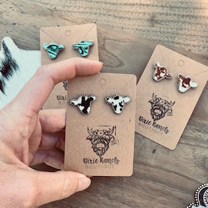 May include: Three pairs of cow-print stud earrings on brown card stock with the text "Dixie Ranches Boutique". The earrings are silver with a black and white cow print design.