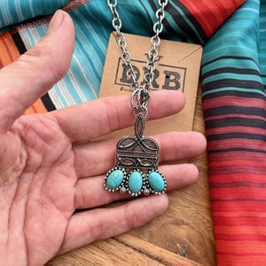 Puede incluir: Collar plateado con un colgante con tres piedras turquesas ovaladas. El colgante tiene un dise&ntilde;o de filigrana ornamentado. El collar se muestra sobre una tela a rayas colorida y una etiqueta de cart&oacute;n marr&oacute;n.