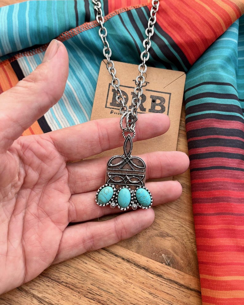 Puede incluir: Collar plateado con un colgante con tres piedras turquesas ovaladas. El colgante tiene un dise&ntilde;o de filigrana ornamentado. El collar se muestra sobre una tela a rayas colorida y una etiqueta de cart&oacute;n marr&oacute;n.