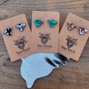 May include: Three pairs of cow-print stud earrings on brown card stock with the text "Dixie Ranches Boutique". The earrings are in black and white, turquoise and white, and brown and white.