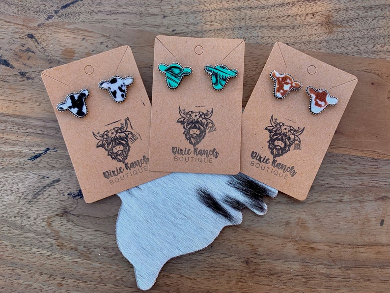 May include: Three pairs of cow-print stud earrings on brown card stock with the text "Dixie Ranches Boutique". The earrings are in black and white, turquoise and white, and brown and white.